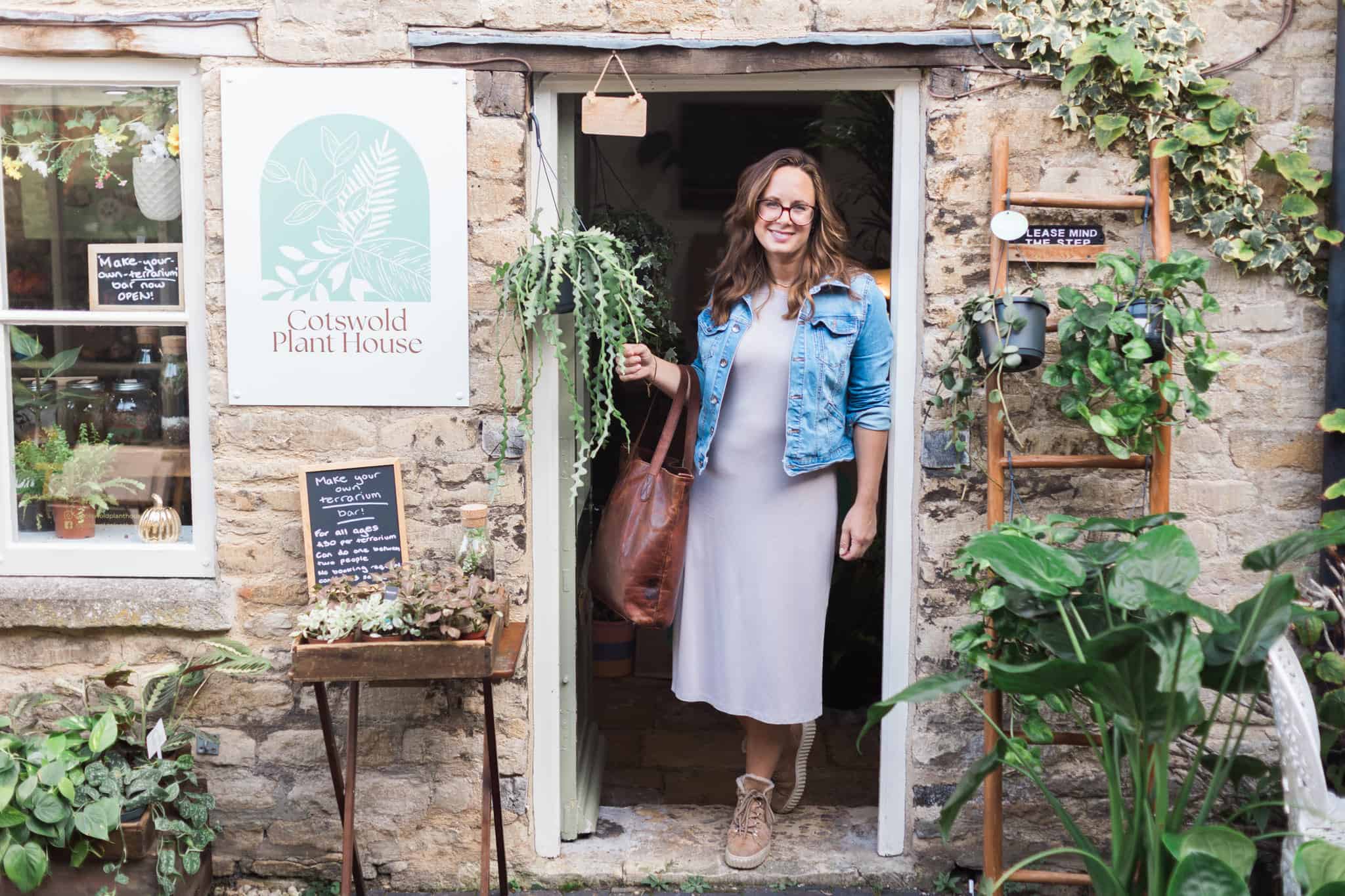 Lady in a neutral dress and denim jacket stands in the entrance to a Cotswold plant shop, smiling at the camera.