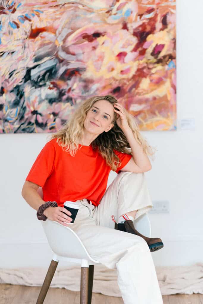 Abstract artist Emma Howell sits in a chair wearing a red t-shirt, in front of one of her colourful large scale paintings