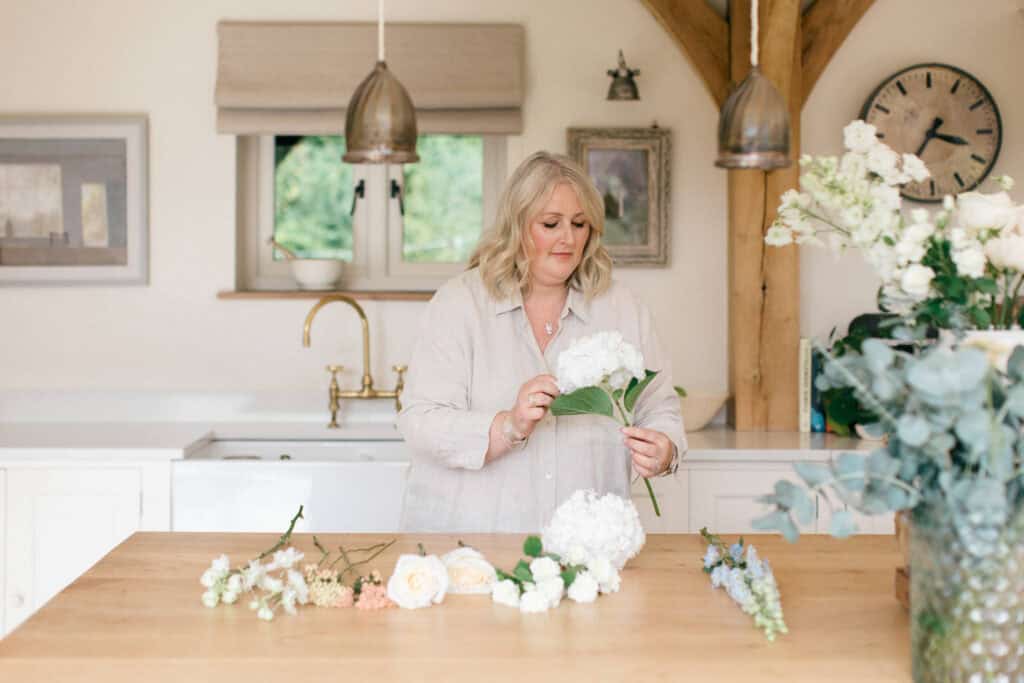 Florist Jo Williams from Issy & Bella at work in a clean spacious kitchen, working with flowers, with stems laid out on the wooden worktop in front of her.